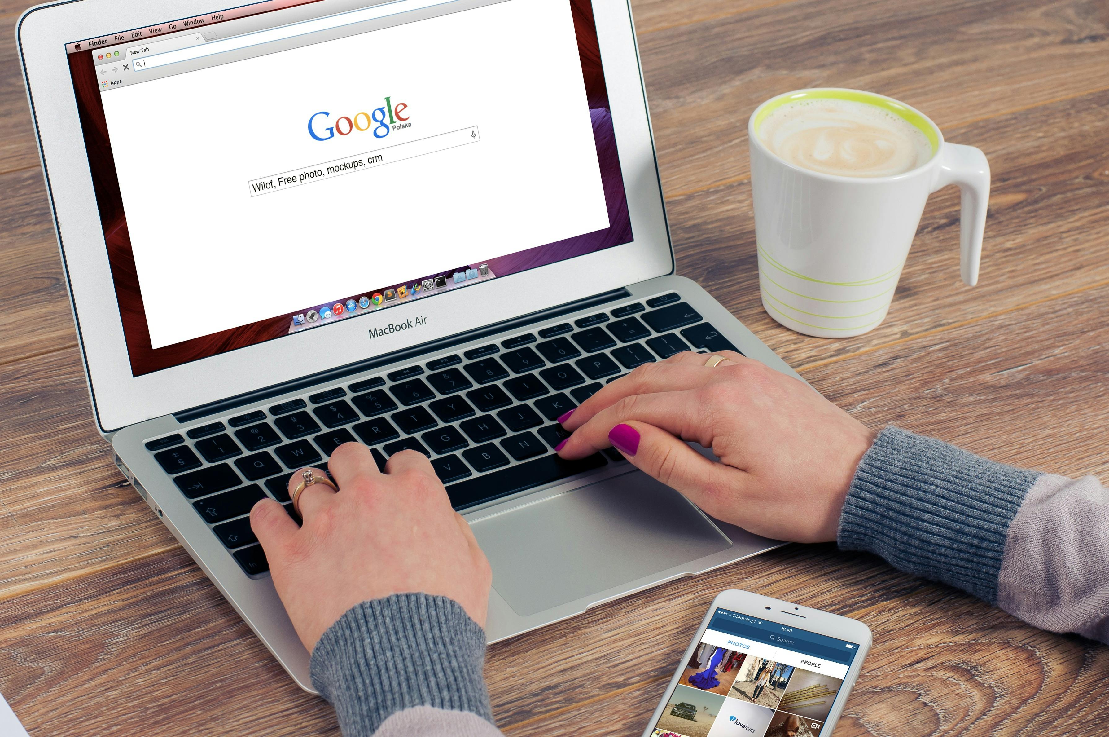 person typing on Google on a laptop, a cell phone in front of them and a coffee to the right of the computer.