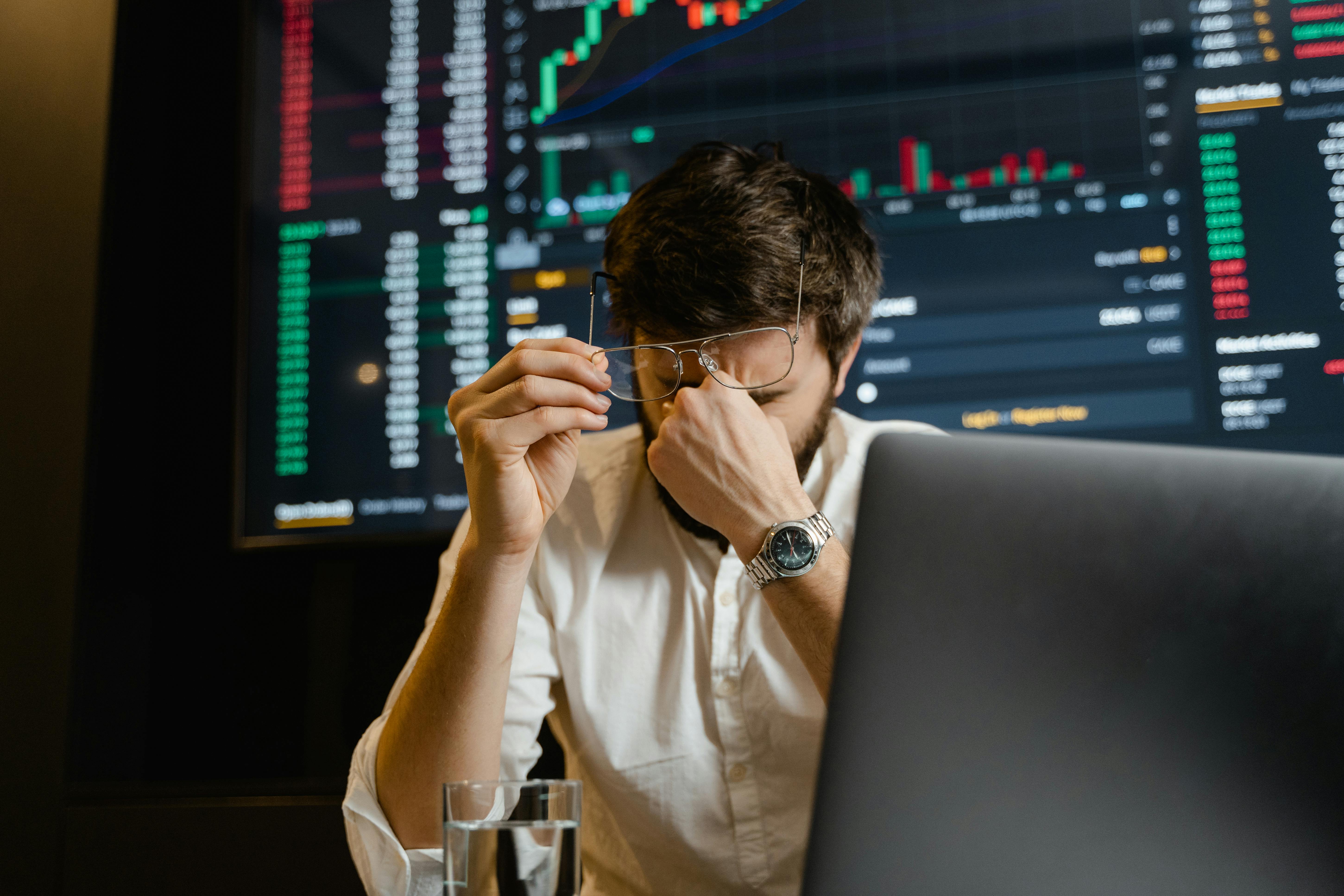 Stressed worker in an office environment. There is a screen with numbers behind him and he's pinching his nose.